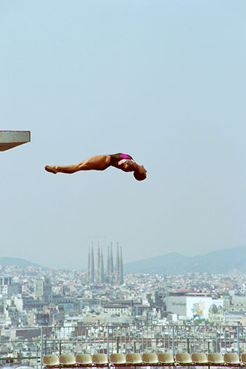 Una saltadora llençant-se des del trampolí de 10 metres de la piscina de Montjuïc rehabilitada per larquitecte Antoni Moragas, amb la ciutat densa al fons.